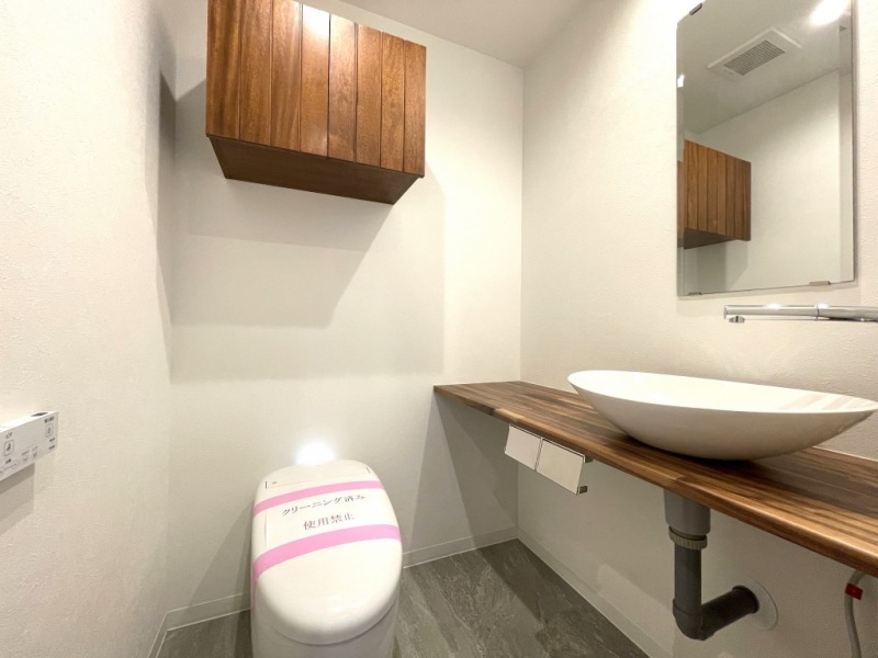 【個室トイレ】タンクレスですっきりとしたデザインのウォシュレット付きトイレです。室内は天然無垢材の温もりを感じる落ち着いた上質な空間です。吊戸棚も設置されており、トイレットペーパーなどの消耗品の収納ができます。ミラー付の便利な専用手洗いスペースも付いています。