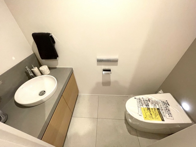 【玄関側トイレ】タンクレスのウォシュレット付きトイレで、室内は清潔感があり落ち着く上質な空間です。吊戸棚も設置されており、トイレットペーパーなどの消耗品の収納ができます。便利な専用手洗いスペースも付いています。