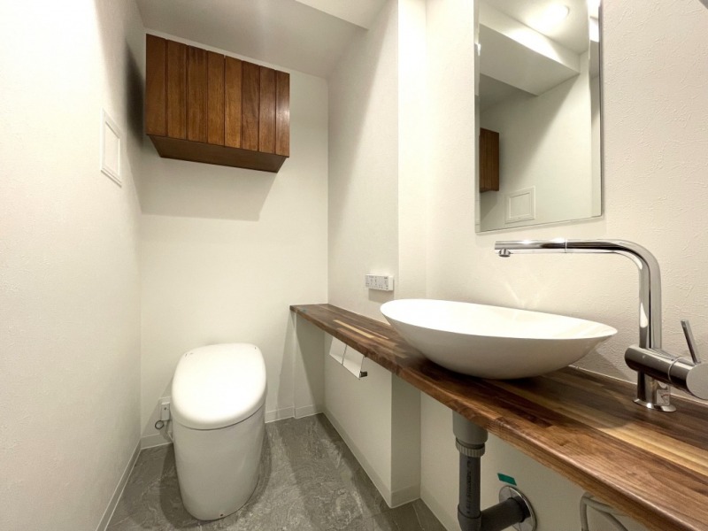 【トイレ】個別のトイレは、こちらもタンクレスのウォシュレット付きトイレです。室内は天然無垢材の温もりを感じる落ち着いた上質な空間です。吊戸棚も設置されており、トイレットペーパーなどの消耗品の収納ができます。ミラー付の便利な専用手洗いスペースが設けられています。