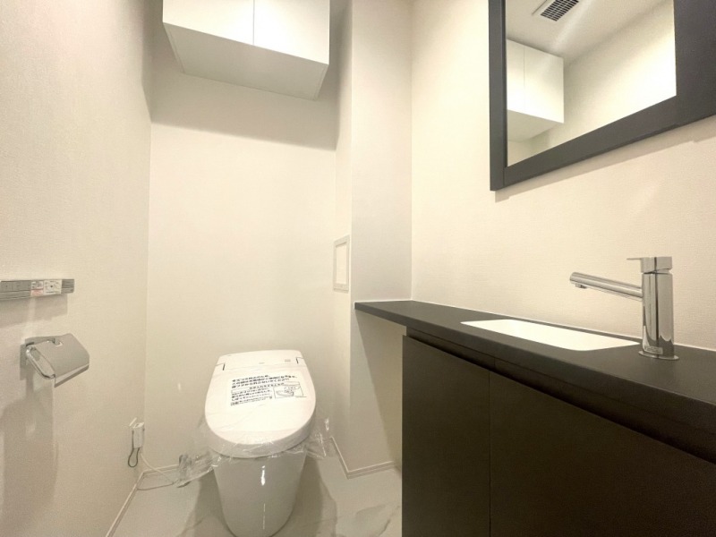 【トイレ】タンクレスの温水洗浄機能付きトイレで、お手入れもしやすくすっきりしています。室内は清潔感があり落ち着く上質な空間です。吊戸棚も設置されており、トイレットペーパーやタオルなどの収納ができます。ミラー付きの便利な専用手洗いスペースも設けられています。