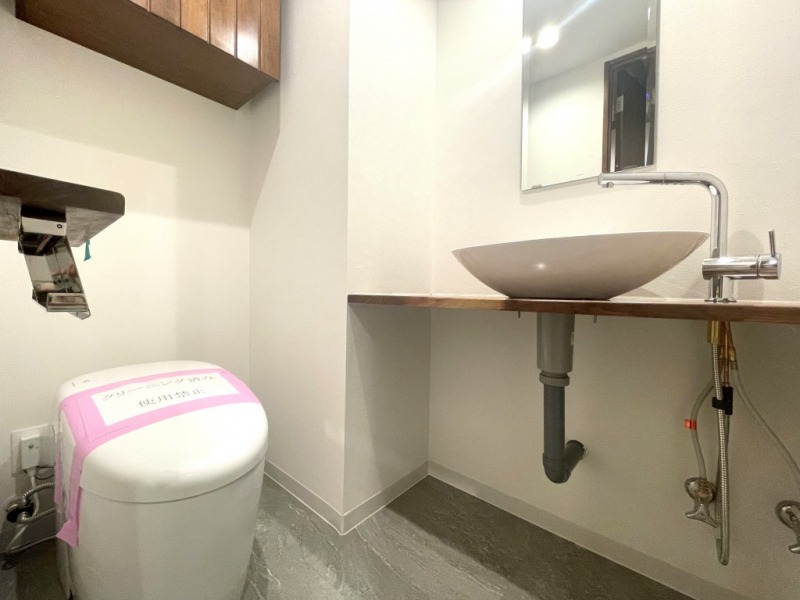 【トイレ】タンクレスですっきりとしたデザインの温水洗浄機能付きトイレです。室内は天然無垢材の温もりを感じる落ち着いた上質な空間です。吊戸棚も設置されており、トイレットペーパーやタオルなどの収納ができます。ミラー付の便利な専用手洗いスペースが設けられています。
