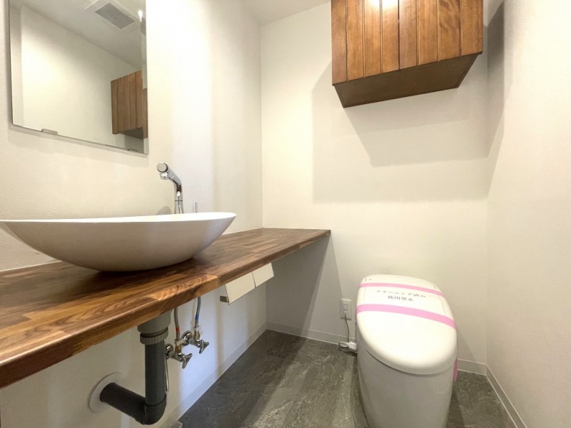 【トイレ】タンクレスですっきりとしたデザインの洗浄機能付きトイレです。室内は天然無垢材の温もりを感じる落ち着いた上質な空間です。吊戸棚も設置されており、トイレットペーパーやタオルなどの収納ができます。ミラー付の便利な専用手洗いスペースが設けられています。