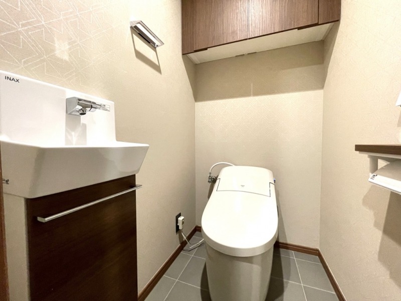 【トイレ】タンクレスの温水洗浄機能付きトイレで、お手入れもしやすくすっきりしています。室内は清潔感があり落ち着く上質な空間です。吊戸棚も設置されており、トイレットペーパーやタオルなどの収納ができます。便利な専用手洗いスペースも設けられています。