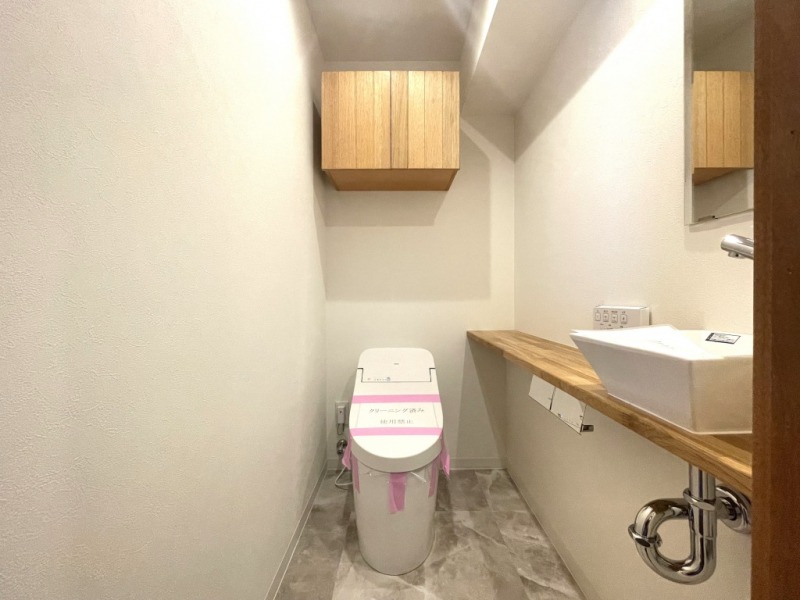 【トイレ】タンクレスですっきりとしたデザインの洗浄機能付きトイレです。室内は天然無垢材の温もりを感じる落ち着いた上質な空間です。吊戸棚も設置されており、トイレットペーパーやタオルなどの収納ができます。ミラー付の便利な専用手洗いスペースが設けられています。