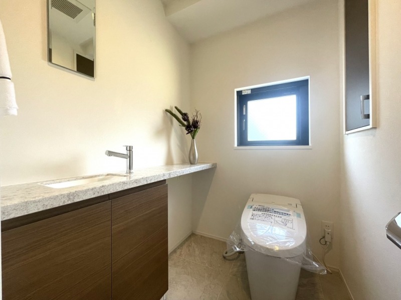 【トイレ】タンクレスの温水洗浄機能付きトイレで、お手入れもしやすくすっきりしています。室内は清潔感があり落ち着く上質な空間です。戸棚も設置されており、トイレットペーパーやタオルなどの収納ができます。カウンターとミラー付きの便利な専用手洗いスペースも設けられています。また窓があるので明るく、こもりがちな臭いも換気ができて快適です。