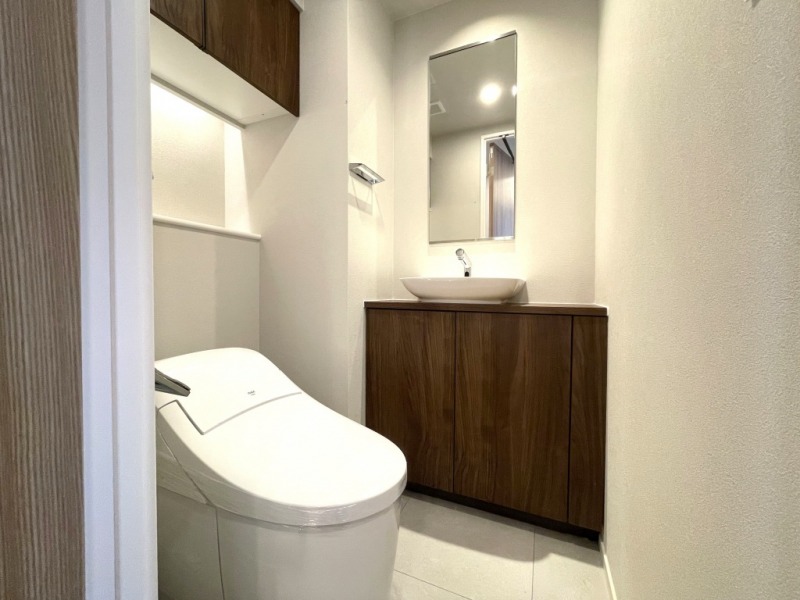 【トイレ】タンクレスですっきりとしたデザインの温水洗浄機能付きトイレです。室内は清潔感があり落ち着く上質な空間です。トイレットペーパーなどが収納できる吊戸棚も設置されています。ミラー付きの便利な専用手洗いスペースも設けられています。