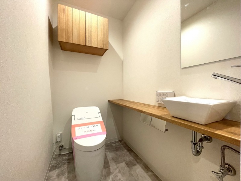 【トイレ】タンクレスですっきりとしたデザインの温水洗浄機能付きトイレです。室内は天然無垢材の温もりを感じる落ち着いた上質な空間です。吊戸棚も設置されており、トイレットペーパーやタオルなどの収納ができます。カウンターやミラー付の便利な専用手洗いスペースが設けられています。