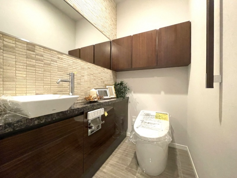【トイレ】タンクレスの温水洗浄機能付きトイレで、お手入れもしやすくすっきりしています。室内は清潔感があり落ち着く上質な空間です。吊戸棚も設置されており、トイレットペーパーやタオルなどの収納ができます。カウンター付きの便利な専用手洗いスペースも設けられています。