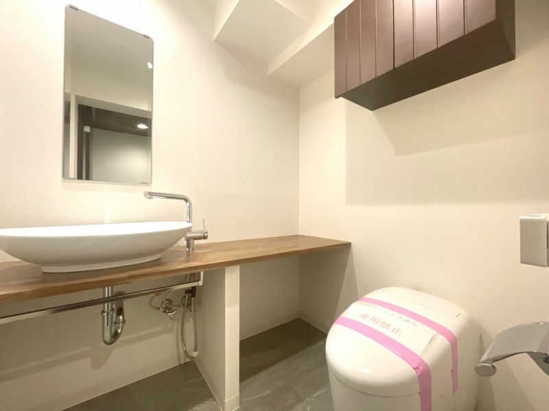 【トイレ】タンクレスですっきりとしたデザインの洗浄機能付きトイレです。室内は天然無垢材の温もりを感じる落ち着いた上質な空間です。吊戸棚も設置されており、トイレットペーパーやタオルなどの収納ができます。ミラー付の便利な専用手洗いスペースが設けられています。
