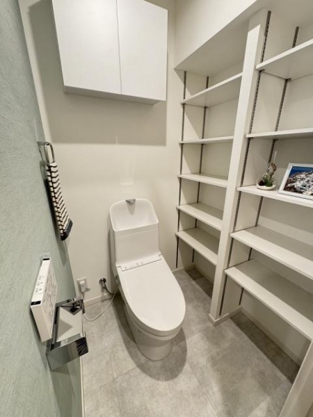 【トイレ】すっきりとしたデザインで清潔感があり、落ち着ける空間です。トイレもうれしい洗浄機能付きです。吊戸棚も設置されており、トイレットペーパーなどが収納できます。多くの可動棚を備えており、飾り棚や本棚として利用できます。本も日焼けをしないので、好適かもしれません。