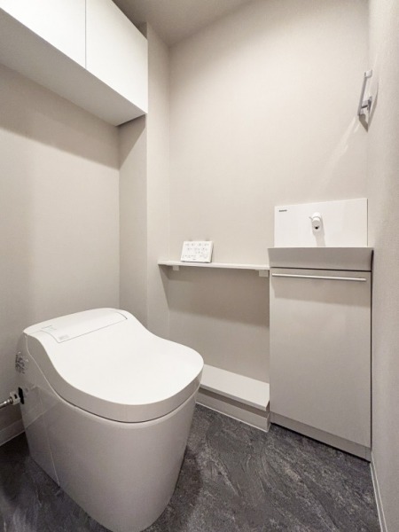 【トイレ】お手入れもしやすいタンクレスで、すっきりとしたデザインの温水洗浄機能付きトイレです。室内は清潔感があり落ち着く上質な空間です。トイレットペーパーなどが収納できる吊戸棚も設置されています。便利な専用手洗いスペースも設けられています。