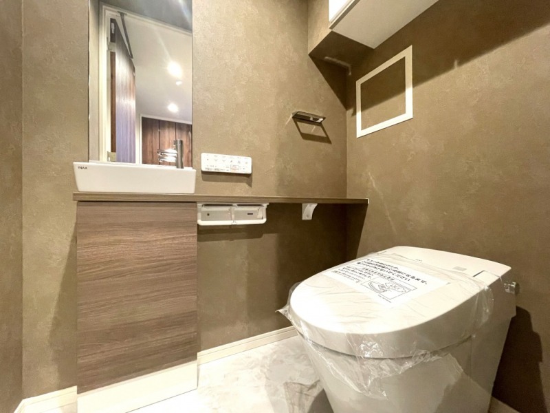 【トイレ】タンクレスですっきりとしたデザインでお手入れもしやすい温水洗浄機能付きトイレです。室内は清潔感があり落ち着く上質な空間です。トイレットペーパーなどが収納できる吊戸棚も設置されています。ミラー付きで便利な専用手洗いスペースも設けられています。