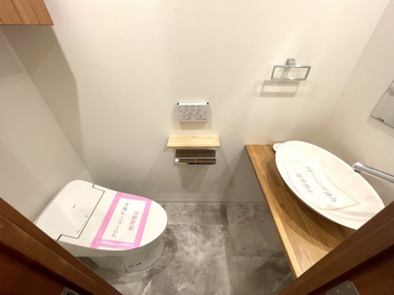 【トイレ】タンクレスですっきりとしたデザインの温水洗浄機能付きトイレです。室内は天然無垢材の温もりを感じる落ち着いた上質な空間です。トイレットペーパーなどが収納できる吊戸棚も設置され、ミラー付の便利な専用手洗いスペースも設けられています。