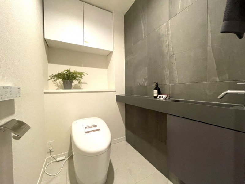 【トイレ】お手入れのしやすいタンクレスで、すっきりとしたデザインの温水洗浄機能付きトイレです。室内はシックなアクセントウォールの落ち着く上質な空間です。トイレットペーパーなどが収納できる吊戸棚も設置されています。カウンター付きの便利な専用手洗いスペースも設けられています。