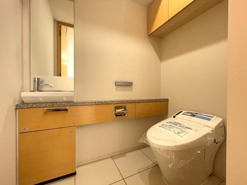 【トイレ】お手入れのしやすいタンクレスで、すっきりとしたデザインの温水洗浄機能付きトイレです。室内は清潔感があり落ち着く上質な空間です。トイレットペーパーなどが収納できる吊戸棚も設置されています。ミラーとカウンター付きの便利な専用手洗いスペースも設けられています。