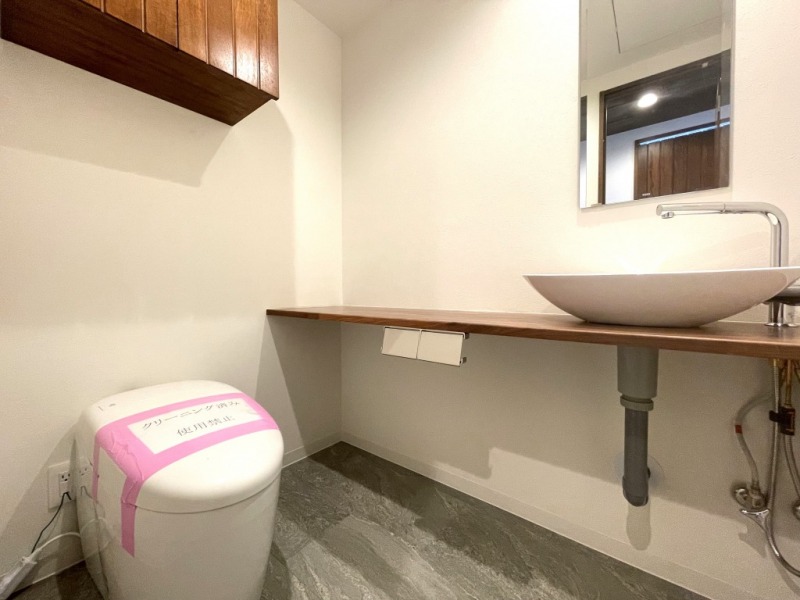 【トイレ】タンクレスですっきりとしたデザインの温水洗浄機能付きトイレです。室内は天然無垢材の温もりを感じる落ち着いた上質な空間です。吊戸棚も設置されており、トイレットペーパーやタオルなどの収納ができます。ミラー付の便利な専用手洗いスペースが設けられています。