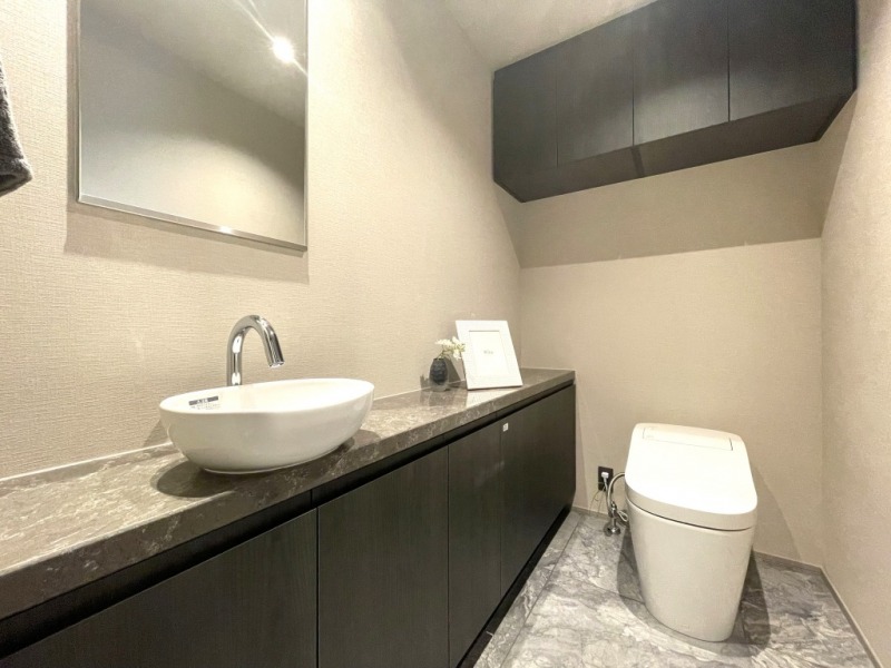 【トイレ】お手入れのしやすいタンクレスで、すっきりとしたデザインの温水洗浄機能付きトイレです。室内は清潔感があり落ち着く上質な空間です。トイレットペーパーなどが収納できる吊戸棚も設置されています。便利な専用手洗いスペースも設けられています。