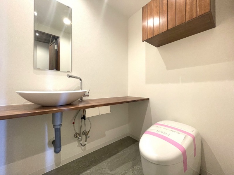 【トイレ】お手入れのしやすいタンクレスで、すっきりとしたデザインの温水洗浄機能付きトイレです。室内は清潔感があり落ち着く上質な空間です。トイレットペーパーなどが収納できる吊戸棚も設置されています。ミラー付きの便利な専用手洗いスペースも設けられています。
