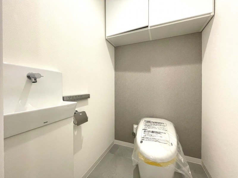 【トイレ】お手入れのしやすいタンクレスで、すっきりとしたデザインの温水洗浄機能付きトイレです。室内は清潔感があり落ち着く上質な空間です。トイレットペーパーなどが収納できる吊戸棚も設置されています。便利な専用手洗いスペースも設けられています。