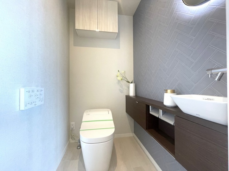 【トイレ】お手入れもしやすいタンクレスで、すっきりとしたデザインの温水洗浄機能付きトイレです。室内は寒色系のアクセントクロスで清潔感があり落ち着く上質な空間です。トイレットペーパーなどが収納できる吊戸棚も設置されています。便利な専用手洗いスペースも設けられています。