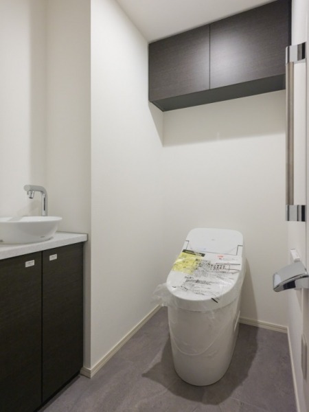 【トイレ】お手入れのしやすいタンクレスで、すっきりとしたデザインの温水洗浄機能付きトイレです。室内は清潔感があり落ち着く上質な空間です。トイレットペーパーなどが収納できる吊戸棚も設置されています。便利な専用手洗いスペースも設けられています。