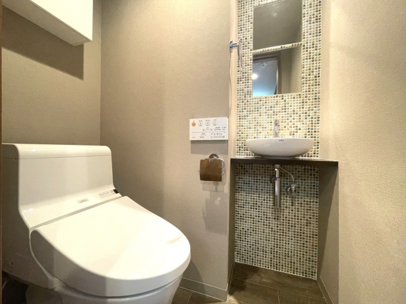 【トイレ】すっきりとしたデザインの温水洗浄機能付きトイレです。室内は清潔感があり落ち着く上質な空間です。トイレットペーパーなどが収納できる吊戸棚も設置されています。タイル貼りのおしゃれな便利な専用手洗いスペースも設けられています。