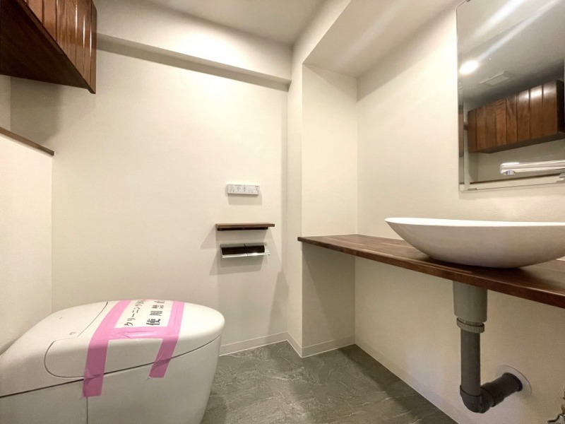 【トイレ】タンクレスですっきりとしたデザインの温水洗浄機能付きトイレです。吊戸棚も設置されており、トイレットペーパーやタオルなどの収納ができます。ミラー付の便利な専用手洗いスペースが設けられています。