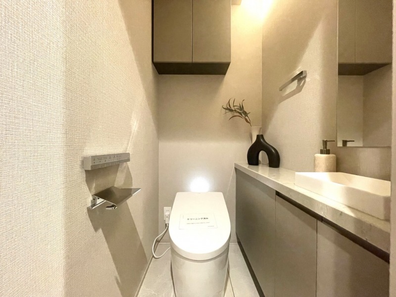 【トイレ】お手入れのしやすいタンクレスで、すっきりとしたデザインの温水洗浄機能付きトイレです。室内は清潔感があり落ち着く上質な空間です。トイレットペーパーなどが収納できる吊戸棚も設置されています。ミラーとカウンター付きの便利な専用手洗いスペースも設けられています。
