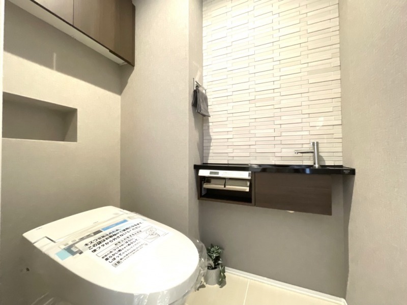 【トイレ】お手入れのしやすいタンクレスで、すっきりとしたデザインの温水洗浄機能付きトイレです。室内は清潔感があり落ち着く上質な空間です。トイレットペーパーなどが収納できる吊戸棚も設置されています。カウンター付きの便利な専用手洗いスペースも設けられています。