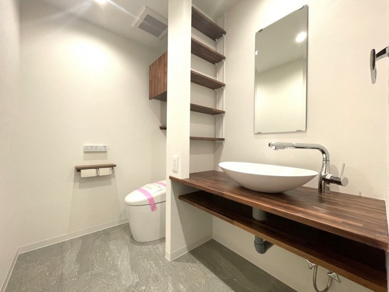 【トイレ】タンクレスですっきりとしたデザインのウォシュレット付きトイレが洗面台奥にあります。吊戸棚も設置されており、トイレットペーパーなどの消耗品の収納ができます。トイレは全部で3か所あります。