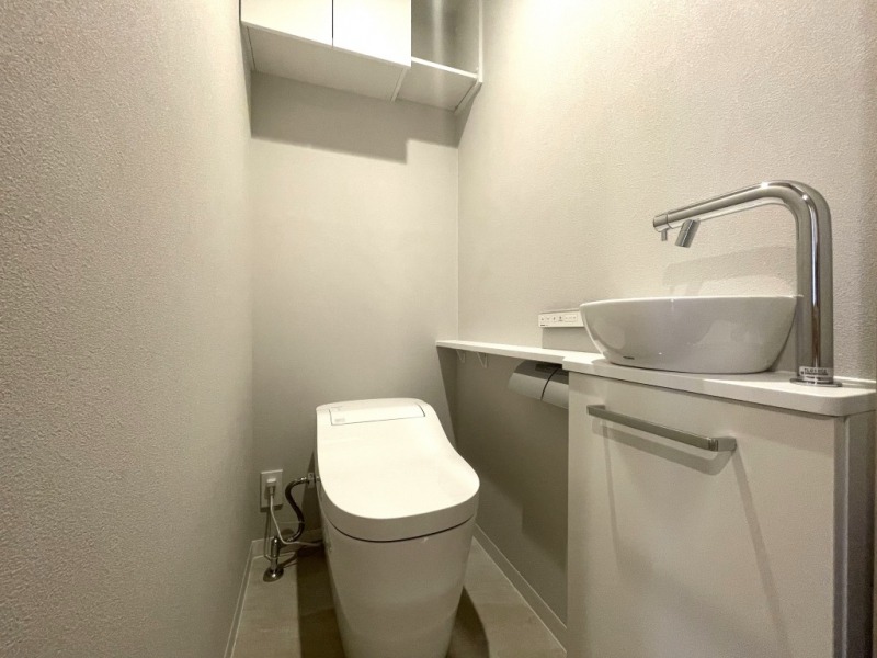 【トイレ】お手入れのしやすいタンクレスで、すっきりとしたデザインの温水洗浄機能付きトイレです。室内は清潔感があり落ち着く上質な空間です。トイレットペーパーなどが収納できる吊戸棚も設置されています。便利な専用手洗いスペースも設けられています。