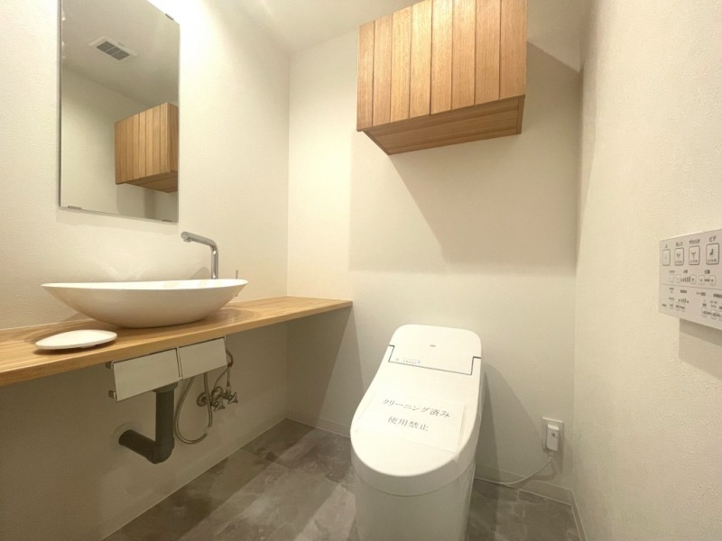 【トイレ：弊社施工事例】タンクレスですっきりとしたデザインの温水洗浄機能付きトイレです。室内は天然木の温もりを感じる落ち着いた上質な空間です。吊戸棚も設置されており、トイレットペーパーやタオルなどの収納ができます。ミラー付の便利な専用手洗いスペースが設けられています。