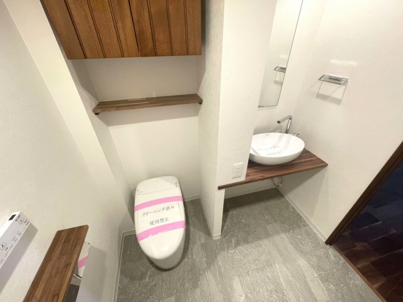 【トイレ】タンクレスですっきりとしたデザインの温水洗浄機能付きトイレです。室内は天然木の温もりを感じる落ち着いた上質な空間です。吊戸棚も設置されており、トイレットペーパーやタオルなどの収納ができます。ミラー付の便利な専用手洗いスペースが設けられています。