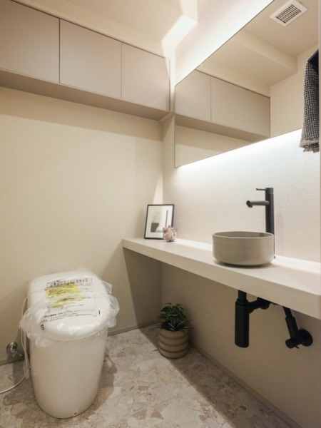 【トイレ】お手入れのしやすいタンクレスで、すっきりとしたデザインの温水洗浄機能付きトイレです。室内は清潔感があり落ち着く上質な空間です。トイレットペーパーなどが収納できる吊戸棚も設置されています。間接照明が灯るミラーやカウンター付きの便利な専用手洗いスペースも設けられています。