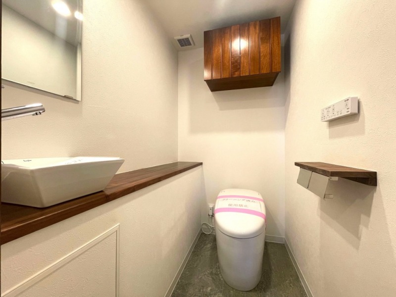 【トイレ】タンクレスですっきりとしたデザインの温水洗浄機能付きトイレです。室内は天然木の温もりを感じる落ち着いた上質な空間。吊戸棚も設置されており、トイレットペーパーやタオルなどの収納ができます。ミラー付の便利な専用手洗いスペースが設けられています。