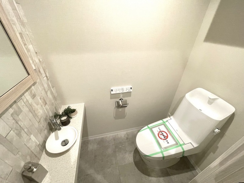 【トイレ】すっきりとしたデザインで清潔感があり、落ち着ける空間です。トイレもうれしい洗浄機能付きです。吊戸棚も設置されており、トイレットペーパーなどが収納できます。ミラーとカウンターの付いた便利な専用手洗いスペースも設けられています。