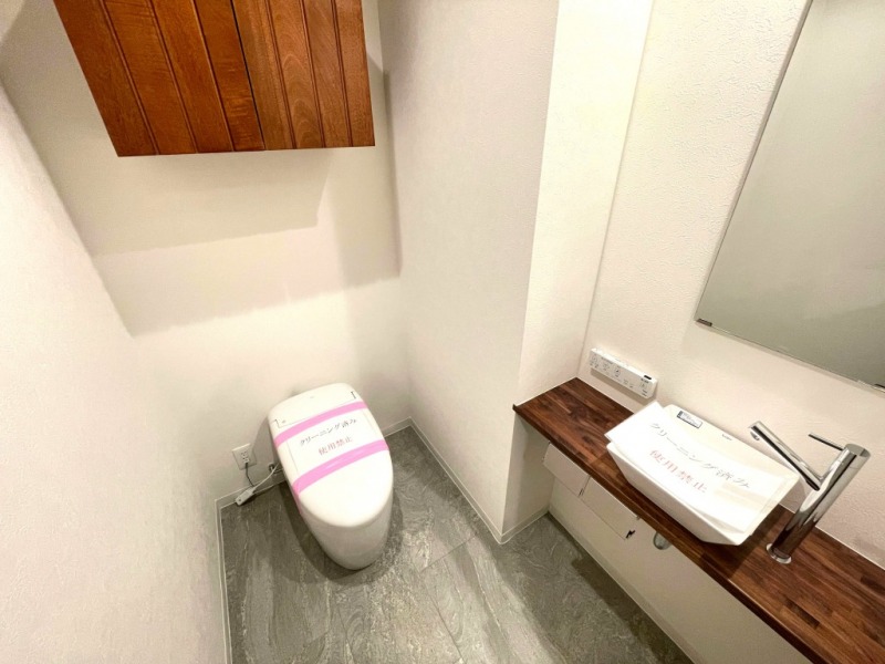 【トイレ】タンクレスですっきりとしたデザインの温水洗浄機能付きトイレです。室内は天然木の温もりを感じる落ち着いた上質な空間。吊戸棚も設置されており、トイレットペーパーやタオルなどの収納ができます。ミラー付の便利な専用手洗いスペースが設けられています。