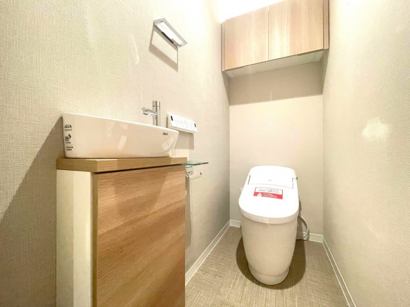 【トイレ】タンクレスですっきりとしたデザインのウォシュレット付きトイレです。室内は木の温もりを感じる落ち着いた上質な空間です。消耗品収納ができる吊り戸棚や便利な専用手洗いスペースが設けられています。