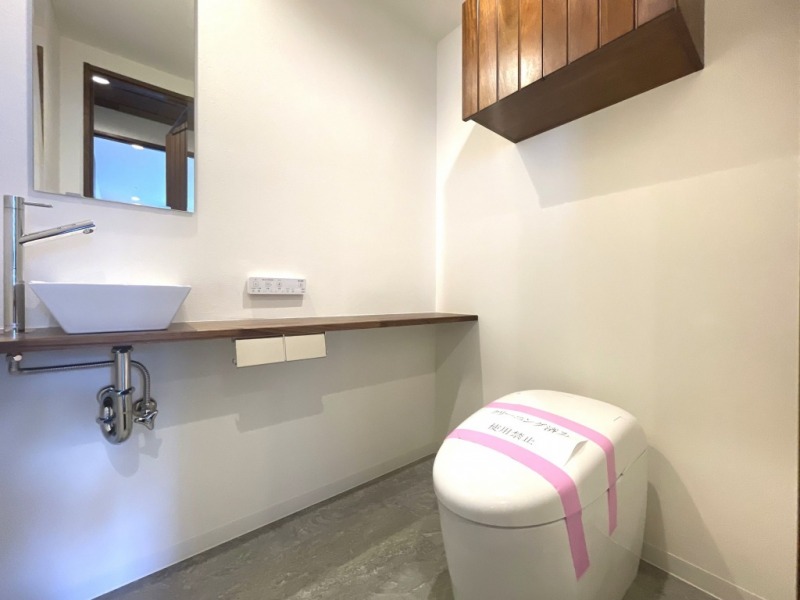 【トイレ:弊社施工事例】タンクレスですっきりとしたデザインの温水洗浄機能付きトイレです。室内は天然木の温もりを感じる落ち着いた上質な空間。吊戸棚も設置されており、トイレットペーパーやタオルなどの収納ができます。ミラー付の便利な専用手洗いスペースが設けられています。