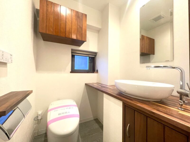 【トイレ】タンクレスですっきりとしたデザインの温水洗浄機能付きトイレです。室内は天然木の温もりを感じる上質な空間。吊戸棚も設置されており、ミラー付の便利な専用手洗いスペースが設けられています。また窓があるので明るく、こもりがちな臭いも換気ができて快適です。