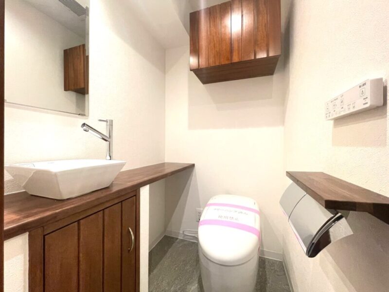 【トイレ】タンクレスですっきりとしたデザインの温水洗浄機能付きトイレです。室内は天然木の温もりを感じる落ち着いた上質な空間。吊戸棚も設置されており、トイレットペーパーやタオルなどの収納ができます。ミラーやカウンター付きの便利な専用手洗いスペースが設けられています。