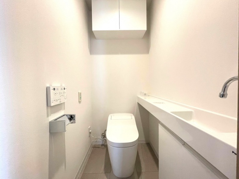 【トイレ】お手入れもしやすいタンクレスのすっきりとしたデザインで温水洗浄機能付きトイレです。室内はホワイトを基調とし、清潔感のある落ち着く空間です。トイレットペーパーなどが収納できる吊戸棚も設置されています。カウンター付きの便利な専用手洗いスペースも設けられています。