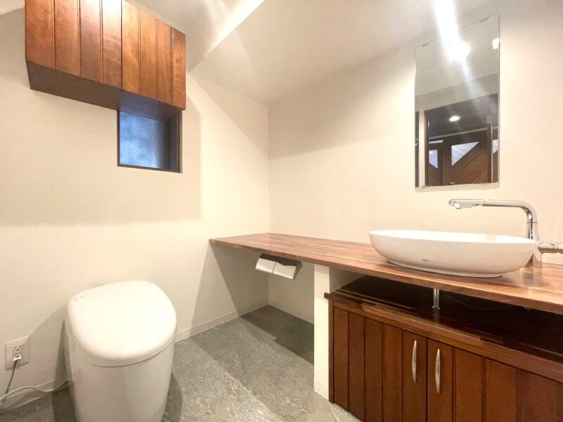 【トイレ】タンクレスですっきりとしたデザインの温水洗浄機能付きトイレです。室内は天然木の温もりを感じる落ち着いた上質な空間。吊戸棚も設置されており、トイレットペーパーやタオルなどの収納ができます。ミラー付の便利な専用手洗いスペースが設けられています。また窓があるので明るく、こもりがちな臭いも換気ができて快適です。