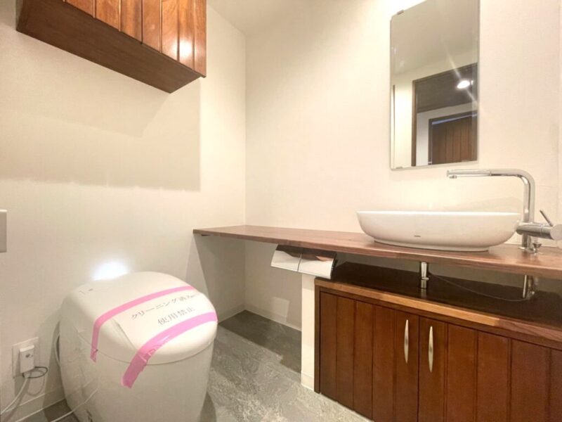 【トイレ】タンクレスですっきりとしたデザインの温水洗浄機能付きトイレです。室内は天然木の温もりを感じる落ち着いた上質な空間。吊戸棚も設置されており、トイレットペーパーやタオルなどの収納ができます。ミラー付の便利な専用手洗いスペースが設けられています。