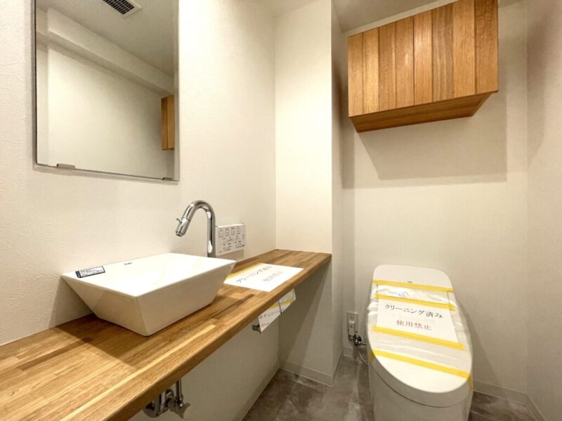 【トイレ:弊社施工事例】タンクレスですっきりとしたデザインの温水洗浄機能付きトイレです。室内は天然木の温もりを感じる落ち着いた上質な空間。吊戸棚も設置されており、トイレットペーパーやタオルなどの収納ができます。カウンターとミラー付の付いた便利な専用手洗いスペースが設けられています。
