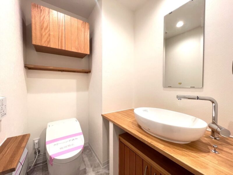 【トイレ：弊社施工事例】タンクレスですっきりとしたデザインの温水洗浄機能付きトイレです。室内は天然木の温もりを感じる落ち着いた上質な空間。吊戸棚も設置されており、トイレットペーパーやタオルなどの収納ができます。ミラー付の便利な専用手洗いスペースが設けられています。
