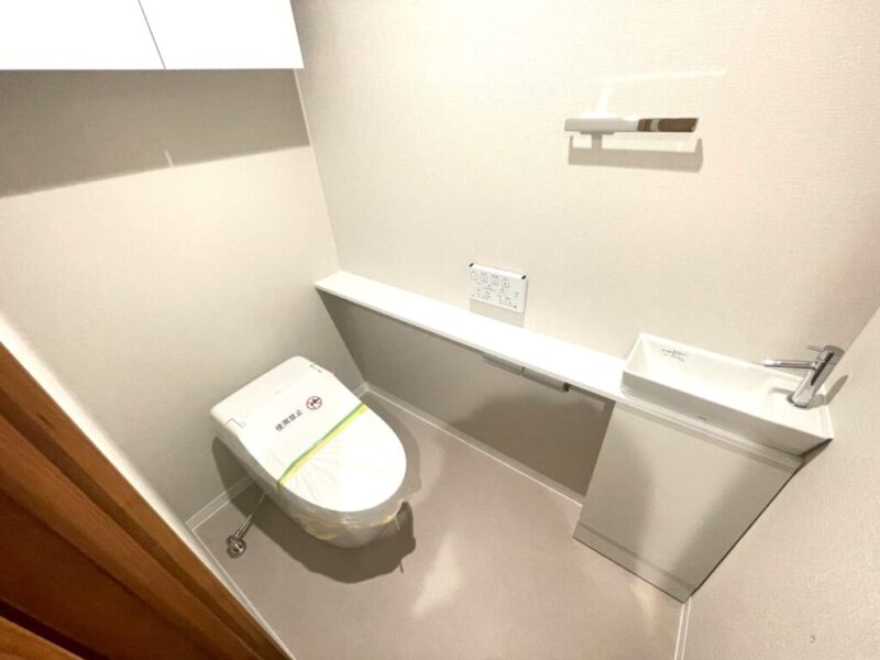 【トイレ】お手入れもしやすいタンクレスのすっきりとしたデザインの温水洗浄機能付きトイレです。室内はホワイトを基調とし、清潔感のある落ち着く空間です。トイレットペーパーなどが収納できる吊戸棚も設置されています。便利な専用手洗いスペースも設けられています。
