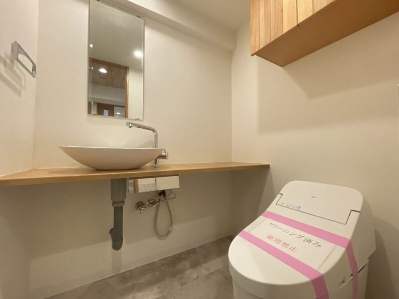 【トイレ:弊社施工事例】タンクレスですっきりとしたデザインの温水洗浄機能付きトイレです。室内は天然木の温もりも感じる落ち着いた上質な空間。吊戸棚も設置されており、トイレットペーパーやタオルなどの収納ができます。ミラーとカウンター付の便利な専用手洗いスペースが設けられています。